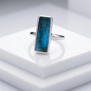 May include: A silver ring featuring a rectangular, vertically oriented, deep blue gemstone set in a smooth metal bezel. The stone displays natural variations in colour and texture, while the band is a simple, polished silver metal wire.