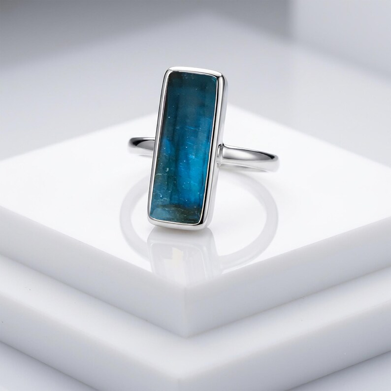 May include: A silver ring featuring a rectangular, vertically oriented, deep blue gemstone set in a smooth metal bezel. The stone displays natural variations in colour and texture, while the band is a simple, polished silver metal wire.