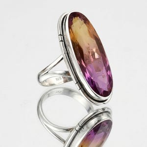 Puede incluir: Anillo grande de forma ovalada con una banda plateada y una piedra preciosa bicolor. La piedra preciosa transita de un tono amarillo dorado en la parte superior a un púrpura intenso en la parte inferior. El anillo está marcado con "925".