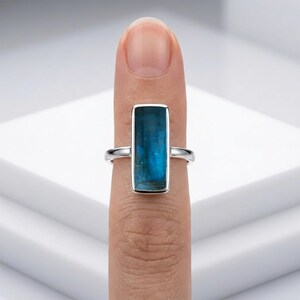May include: A silver ring featuring a rectangular, vertically oriented blue gemstone set in a smooth metal bezel. The band is thin and polished, showcasing the deep teal-blue colour and natural internal inclusions of the stone clearly.