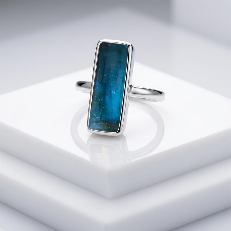 May include: A silver ring featuring a rectangular, vertically oriented, deep blue gemstone set in a smooth metal bezel. The stone displays natural variations in colour and texture, while the band is a simple, polished silver metal wire.
