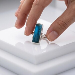 May include: A silver ring featuring a rectangular, vertically oriented blue gemstone set in a smooth metal bezel. The stone displays a deep, translucent teal hue with natural internal inclusions, mounted on a simple, polished silver band.
