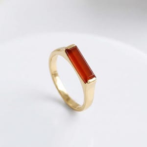 Puede incluir: Un anillo de oro con una piedra preciosa rectangular de color rojo anaranjado. El anillo tiene un diseño simple y elegante con un acabado pulido. La piedra preciosa está engastada en la banda de oro. Joyería.