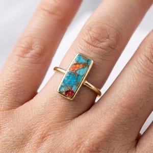 Puede incluir: Un anillo de oro con una piedra rectangular de turquesa, con inclusiones naranjas y rojas. El anillo tiene una fina banda de oro pulido. La piedra está engastada en un bisel de oro, creando un diseño moderno. El anillo se muestra en un dedo.