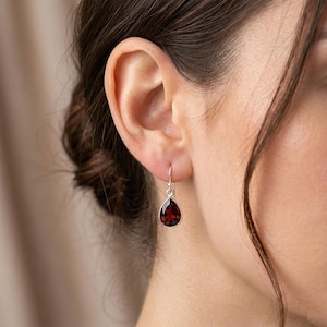 May include: A silver drop earring featuring a faceted, pear-shaped deep red garnet gemstone. The stone is set in a polished silver bezel and hangs from a simple silver hook wire, creating a classic and elegant piece of jewellery for the ear.
