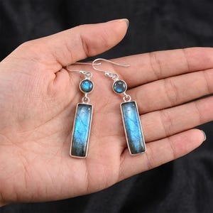 Puede incluir: Pendientes de plata con una piedra rectangular azul y una piedra redonda más pequeña encima. Las piedras tienen tonos azules y turquesas, engastadas en metal plateado. Los pendientes se muestran en una mano.
