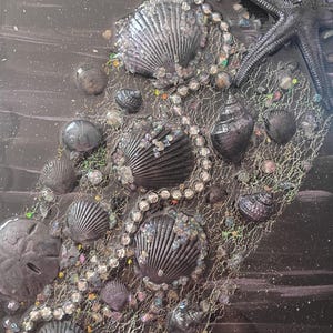 May include: A textured artwork in dark tones, featuring seashells, a starfish, and beads. The shells are grey and silver, with a net-like structure and iridescent highlights. The piece evokes a coastal or nautical theme.