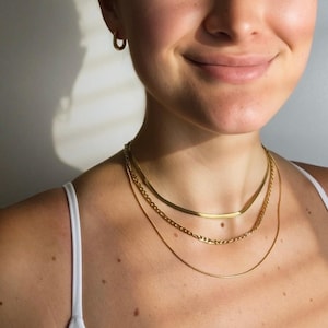 May include: A set of three layered gold-tone necklaces featuring a flat herringbone chain, a medium-weight figaro chain, and a thin snake chain, accompanied by a single small gold-tone hoop earring worn in the earlobe.