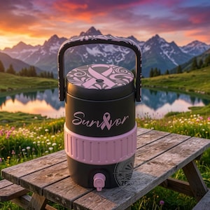 Puede incluir: Un enfriador de bebidas negro mate con un asa rosa y una banda central texturizada rosa. La tapa presenta un diseño de cinta rosa y el frente muestra la palabra Survivor en fuente cursiva con una cinta rosa reemplazando la letra v.