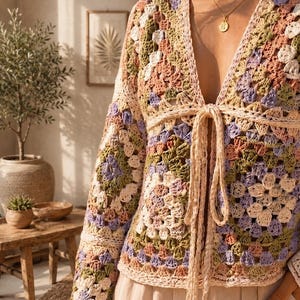 May include: A long-sleeved crochet cardigan featuring a multi-colored granny square pattern in shades of sage green, dusty rose, lavender, and cream. The garment has a deep V-neckline and a front tie closure made of matching cream-colored yarn.