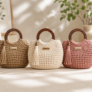 Può includere: Tre borse a mano all'uncinetto nei colori beige, crema e rosa antico. Ogni borsa presenta un manico rotondo in legno scuro, una nappa abbinata attaccata al lato e una piccola targhetta metallica rettangolare al centro con la scritta Handmade.