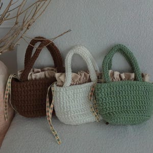 Könnte beinhalten: Drei kleine Häkeltaschen in Dunkelbraun, Cremeweiß und Salbeigrün. Jede Tasche verfügt über einen dicken gehäkelten Henkel, einen gerafften beigen Stoff-Kordelzug und dekorative Bänder mit einem kleinen Blumenmuster an den Seiten.