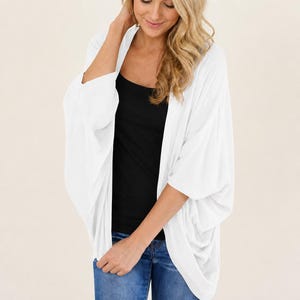 May include: A white, open-front cardigan with three-quarter sleeves. The lightweight fabric drapes loosely. The cardigan is worn over a black tank top and blue jeans. The sleeves are wide and the overall style is casual and relaxed.