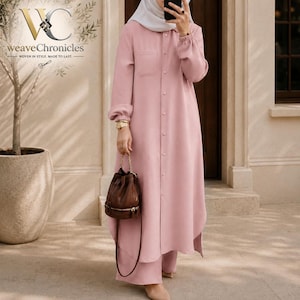 May include: A long-sleeved, button-down shirt dress in a solid dusty pink color, featuring a chest pocket, a classic collar, and a curved hemline. The garment is paired with matching wide-leg trousers and a brown leather bucket bag with gold hardware.