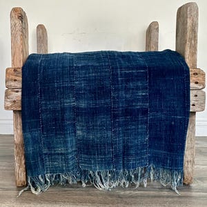 May include: A rectangular, deep indigo blue textile with a visible woven texture and vertical stitched seams, featuring frayed, uneven fringe along the bottom edge, draped over a rustic, light brown wooden stool with four vertical posts.