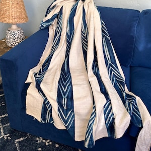 May include: A cream-colored throw blanket with vertical stripes of indigo blue, featuring a repeating chevron pattern. The blanket is draped over a deep blue sofa. A woven lamp with a beige shade sits on a wooden side table.