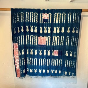 May include: A blue and white patterned textile wall hanging with pink fabric patches sewn on. The textile has a fringed edge.