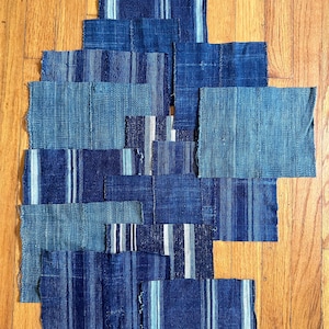 May include: An assortment of indigo blue fabric patches with varying textures and stripe patterns. Some patches feature white or light blue stripes. The patches are arranged on a wooden surface, showcasing the different shades of blue.