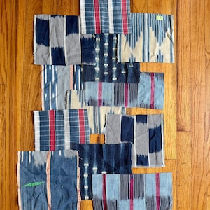 May include: A collection of rectangular fabric swatches in various patterns and colors. The swatches feature designs in blue, red, white, and beige. Some have striped or checkered patterns, while others have more intricate designs. The fabrics are arranged on a wooden surface.