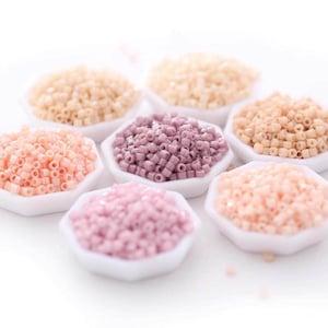 May include: A collection of small, cylindrical seed beads arranged in seven white hexagonal dishes. The beads are presented in various soft pastel shades, including light pink, dusty rose, cream, and beige, each featuring a central hole.