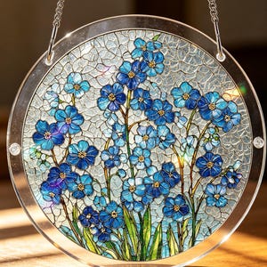 Può includere: Una finestra rotonda in vetro colorato con un motivo floreale di fiori blu non ti scordar di me. I fiori hanno centri gialli e foglie verdi, su uno sfondo azzurro chiaro strutturato. Una catena d'argento è attaccata per appenderla.