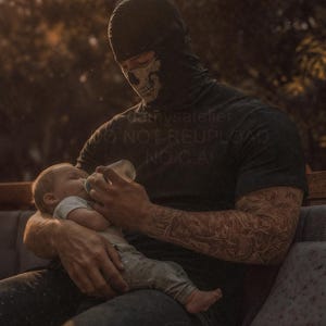Puede incluir: Una persona con un pasamontañas negro con diseño de calavera alimenta a un bebé con un biberón. La persona lleva una camiseta negra con texto. La persona tiene un tatuaje de manga completa en el brazo.
