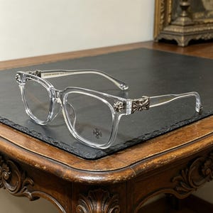 May include: Clear rectangular eyeglasses with silver-coloured decorative accents on the temples and hinges. The lenses are clear, and the frame is transparent. The glasses are resting on a dark surface.