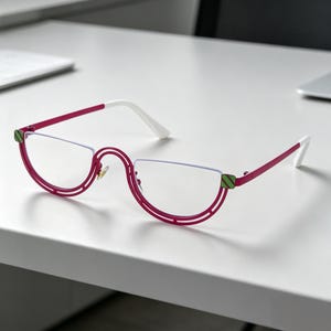 May include: A pair of stylish eyeglasses with a unique half-rim design. The frames are a vibrant magenta color, complemented by white temples and green accents. The lenses are clear, and the glasses are set against a white surface.