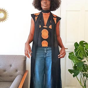 May include: A black sleeveless top and long vest with orange circular patterns. The vest has long fringe. The model is wearing blue jeans and a black choker. A gold mirror is on the wall.