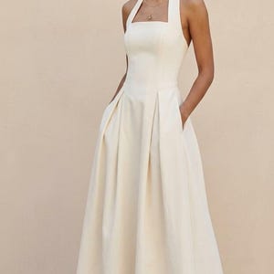 May include: A floor-length cream-colored halter dress featuring a square neckline, a fitted bodice, and a full, pleated skirt with side pockets. The garment is sleeveless with wide straps that secure behind the neck, creating an open back design.