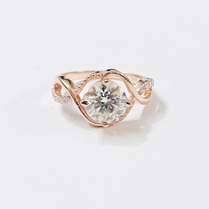 Könnte beinhalten: Ein Ring aus Roségold mit einem zentralen, rund geschliffenen, klaren Stein in einer gedrehten, perlenartigen Fassung. Die Ringschiene teilt sich zu beiden Seiten in zwei zarte, ineinander verschlungene Arme mit kleinen, funkelnden Steinen.