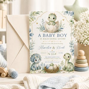 May include: A baby shower invitation card featuring a green dinosaur hatching from an egg, two other dinosaurs, and a nest of eggs. The text reads: A BABY BOY IS HATCHING SOON! Please join us for a baby shower honouring Amelia & Noah, Saturday, October 18, 2:30 PM.