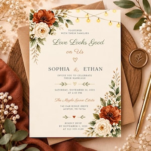 May include: A wedding invitation card featuring cream-colored paper with dark red and white floral arrangements in the corners, string lights at the top, and black text reading Love Looks Good on Us, Sophia and Ethan, and event details.
