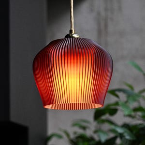 Könnte beinhalten: Eine Pendelleuchte mit einem tiefroten, gerippten Glasschirm in glockenförmiger Silhouette. Der Schirm wird von innen beleuchtet und strahlt ein warmes Licht aus; er ist an einem hellen Kabel befestigt, das in einer messingfarbenen Metallkappe endet.