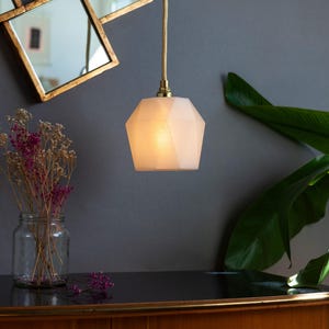 Può includere: Una lampada a sospensione geometrica bianca, con un design sfaccettato, emette una luce calda. È sospesa a un cavo dorato. Sono visibili anche uno specchio dorato e un vaso di vetro con fiori secchi.