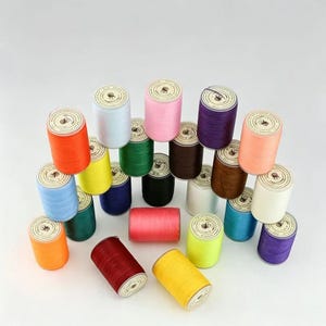 May include: A collection of colorful thread spools in various shades, including orange, yellow, blue, green, pink, and purple. The spools are arranged in a stacked formation, showcasing the diverse color palette of the thread.