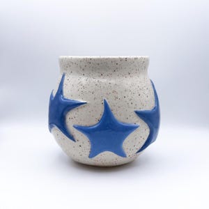 May include: A round ceramic mug with a speckled off-white glaze featuring three raised, glossy blue star shapes applied to the exterior surface. The vessel has a wide, curved body and a smooth, rounded rim, showcasing a handcrafted pottery aesthetic.