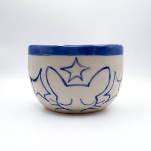 May include: A small, round ceramic bowl with a cream-colored base and a thick blue rim. The exterior features hand-painted blue line art depicting a stylized sleeping cat face with closed eyes and whiskers, surrounded by several five-pointed stars.