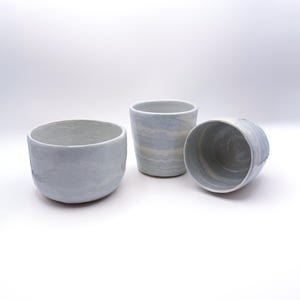 Puede incluir: Tres recipientes de cerámica con un acabado jaspeado en azul claro y blanco. Un recipiente es un cuenco ancho y poco profundo, mientras que los otros dos son tazas cilíndricas más altas. Las superficies presentan un patrón ondulado suave.