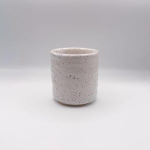 Puede incluir: Una taza cilíndrica de cerámica con un esmalte blanquecino moteado y una superficie sutilmente texturizada. El recipiente tiene un borde recto y una base ligeramente cónica, mostrando un diseño simple y funcional con motas oscuras.