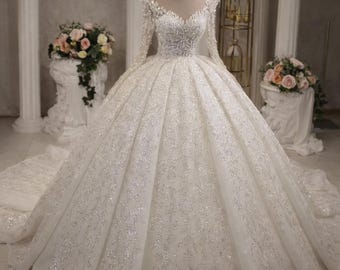 May include: A floor-length white ball gown featuring long sleeves, a sweetheart neckline, and a full, voluminous skirt. The entire dress is heavily embellished with intricate silver beadwork, sequins, and embroidery, paired with a matching tiara.