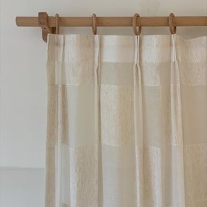 May include: Off-white sheer curtains with a textured, striped pattern suspended from a wooden rod with wooden rings. The curtains have a light and airy appearance, suitable for a living room or bedroom.