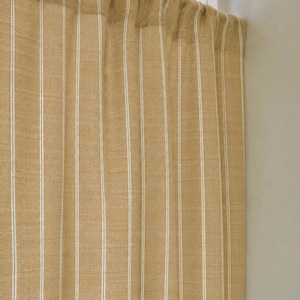 May include: Tan and white striped curtain panel. The curtain has vertical white stripes against a tan background. The fabric appears to be a woven material. The curtain is hanging in front of a window.