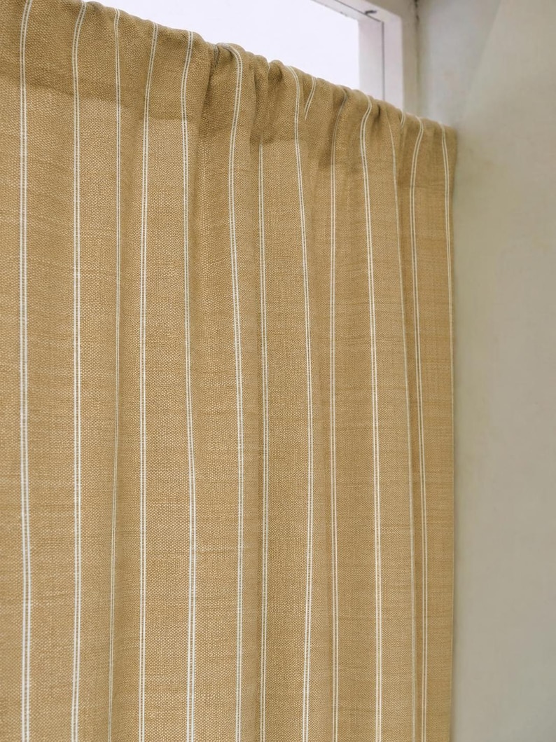 May include: Tan and white striped curtain panel. The curtain has vertical white stripes against a tan background. The fabric appears to be a woven material. The curtain is hanging in front of a window.