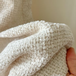 May include: A piece of off-white fabric featuring a dense, textured, and raised grid-like weave. The material has a soft, plush appearance with a consistent light cream color throughout, showcasing the intricate interlacing of the fibers.