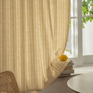 May include: Yellow and white striped curtain hanging in front of a window. A lemon sits on a stack of books on the windowsill. A woven chair and a yellow pillow are in the foreground.