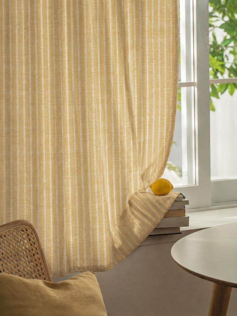 May include: Yellow and white striped curtain hanging in front of a window. A lemon sits on a stack of books on the windowsill. A woven chair and a yellow pillow are in the foreground.