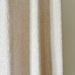 May include: A cream-colored curtain featuring a dense, textured weave with a subtle grid-like pattern. The fabric hangs in vertical folds, creating soft shadows and highlights across the surface, showcasing a consistent, tactile, and woven appearance.
