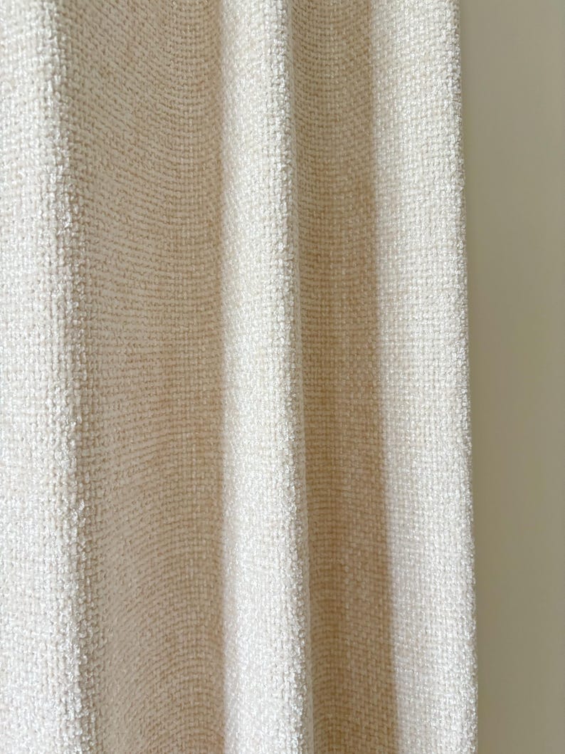 May include: A cream-colored curtain featuring a dense, textured weave with a subtle grid-like pattern. The fabric hangs in vertical folds, creating soft shadows and highlights across the surface, showcasing a consistent, tactile, and woven appearance.