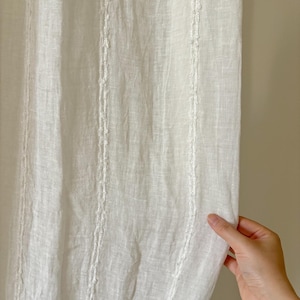 Puede incluir: Un panel de cortina blanco semitransparente con un patrón de rayas verticales y detalles de bordado en relieve. La tela presenta un tejido visible similar al lino, creando una apariencia ligera y aireada con sutiles líneas verticales a lo largo.